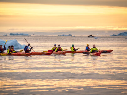 Shows waters in Ilulissat, Greenland, where a group of people are kayaking in between icebergs at Disko Bay, north of the Artic Circle near Ilulissat Icefjord at sunset.