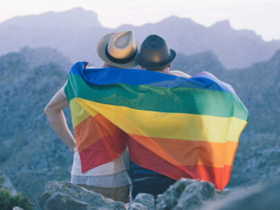Couple sits up in the mountains looking out over the scenery with a LGBTQ+ pride flag around them.