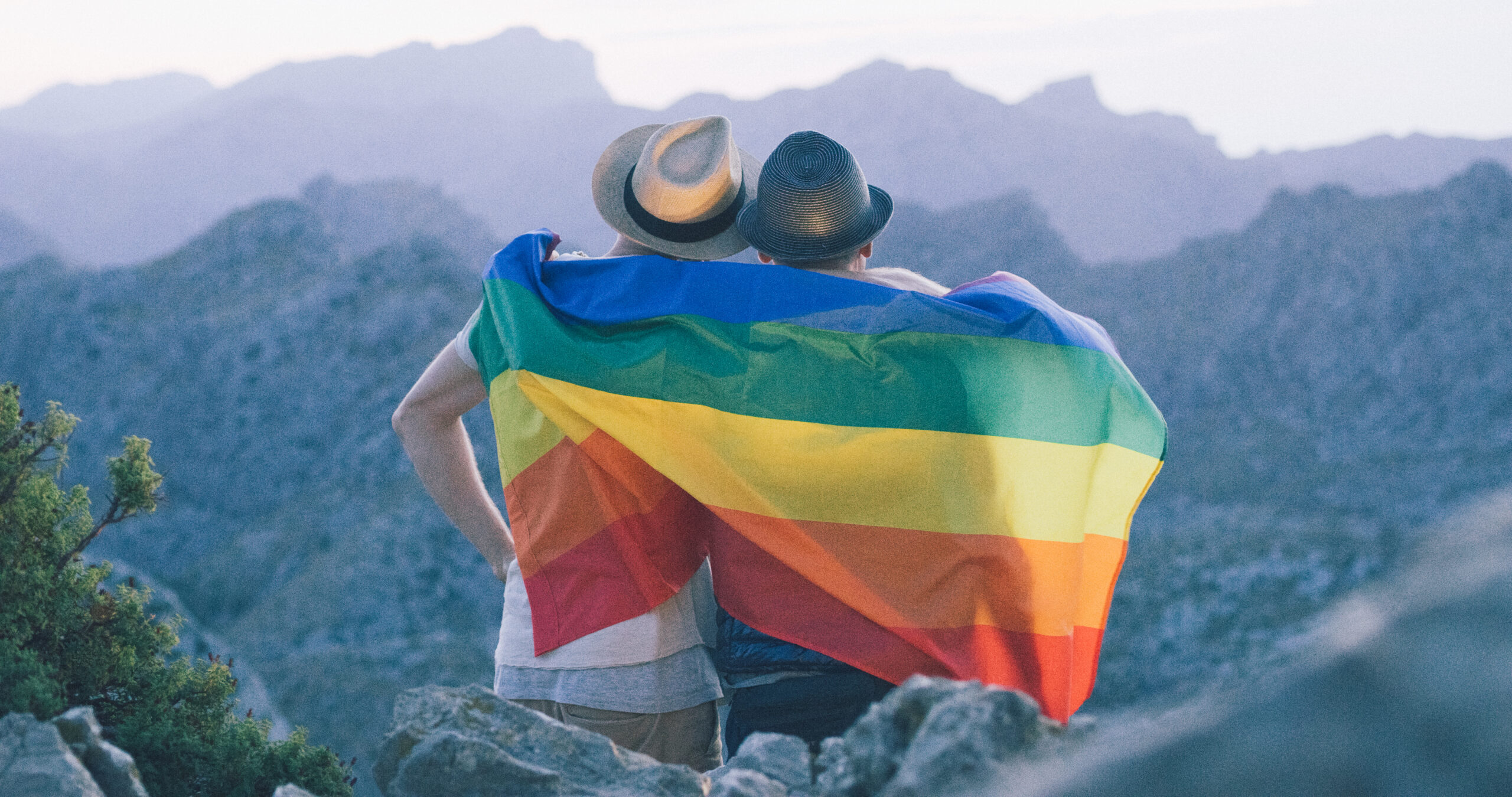 Couple sits up in the mountains looking out over the scenery with a LGBTQ+ pride flag around them.